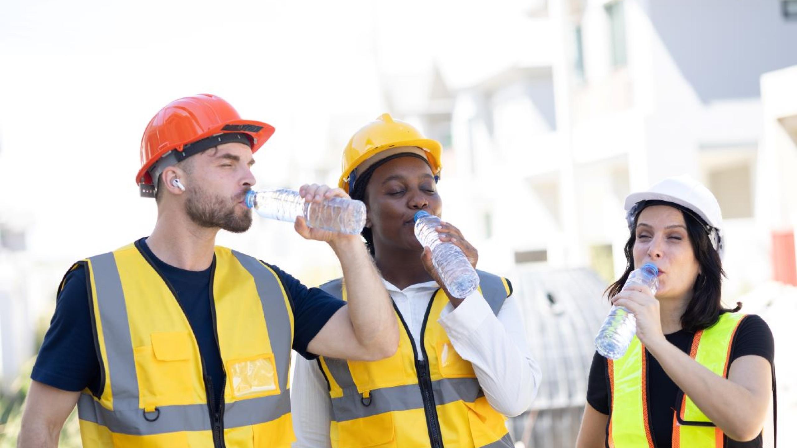 Drie bouwprofessionals op een zonnige werf drinken water.