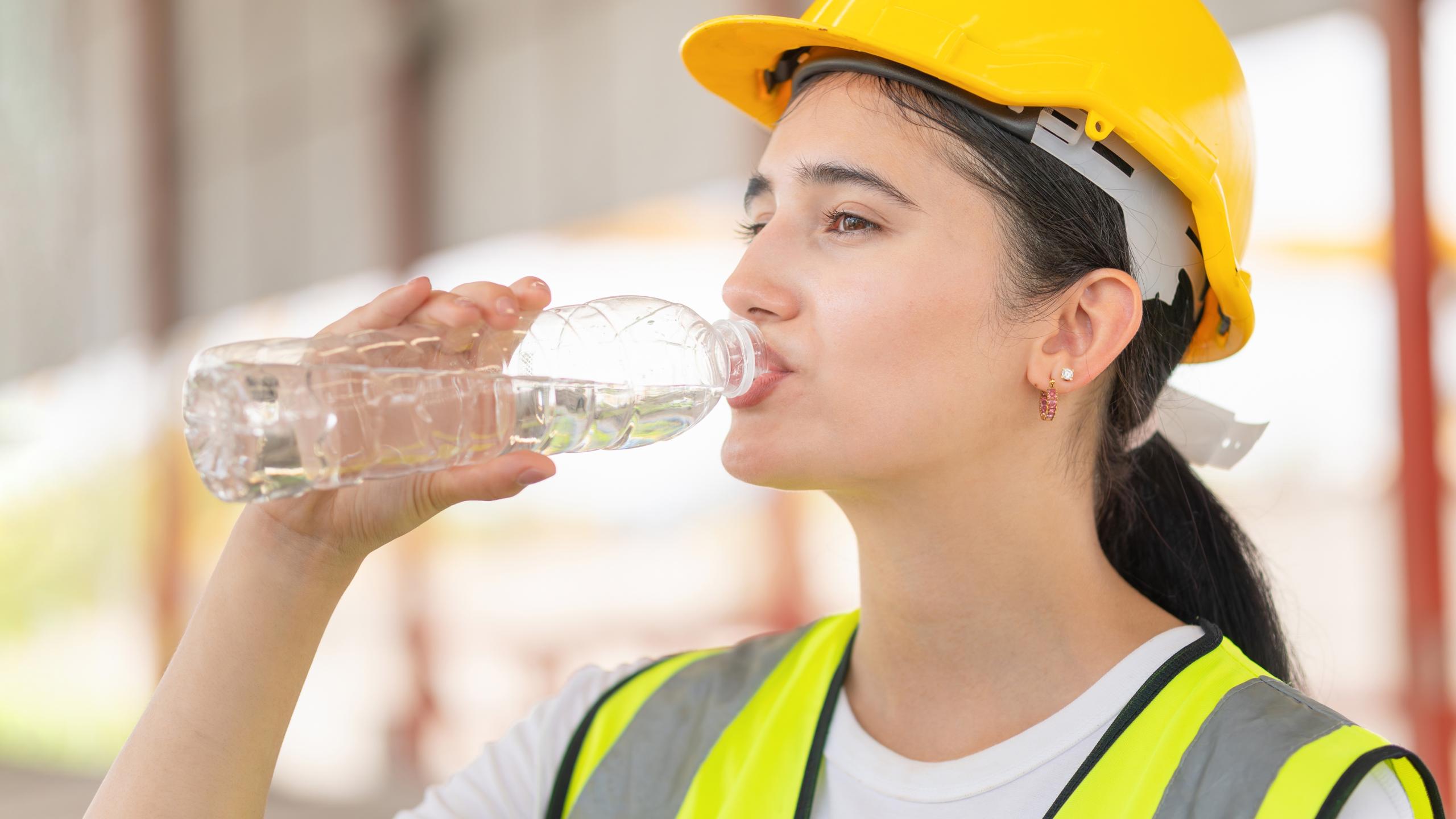 Jonge vrouw met gele helm en fluohesje drink water op bouwplaats.