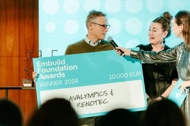 Man met grote cheque in de handen geeft op podium uitleg aan presentator.